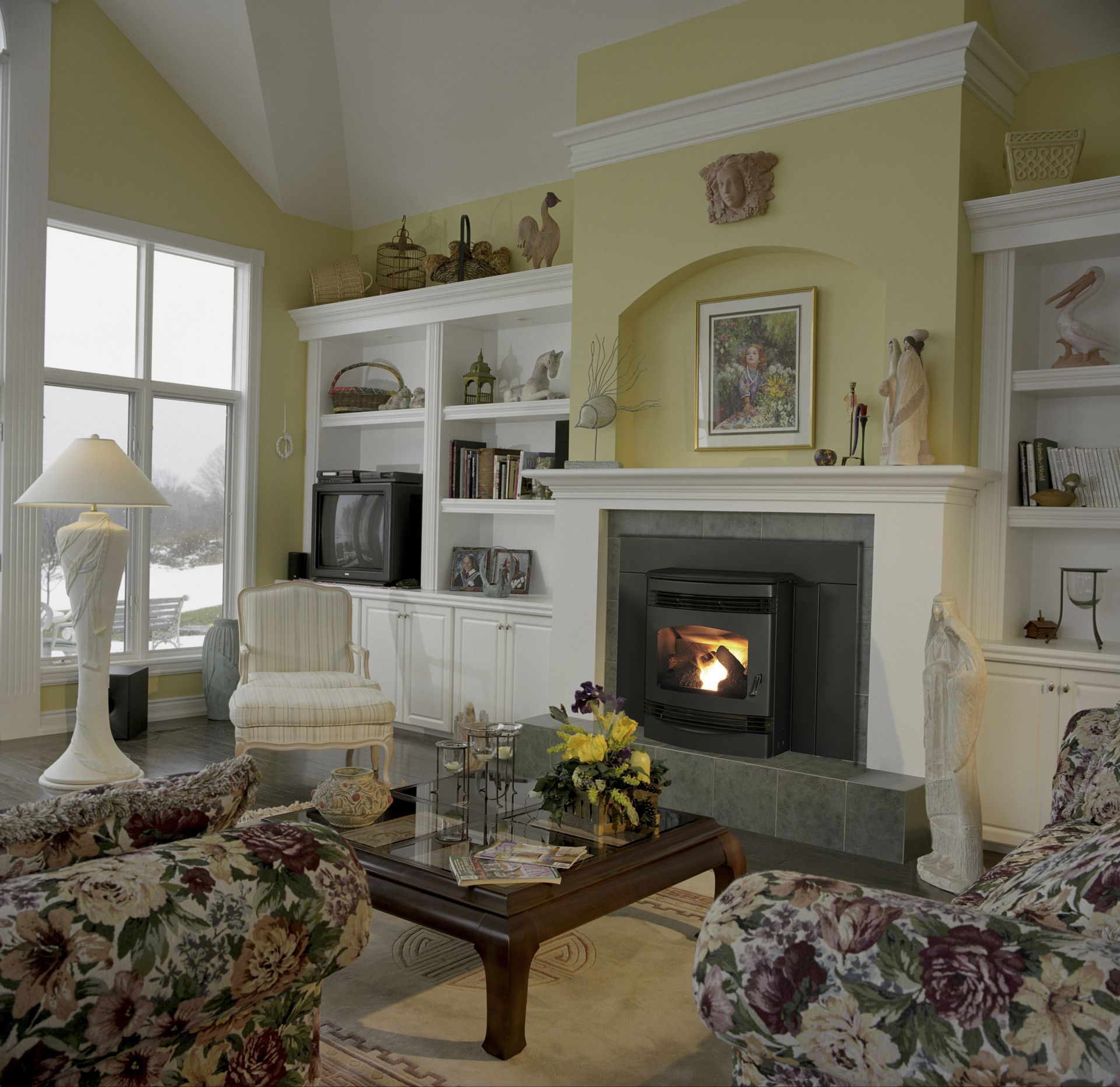 Cozy living room with fireplace, floral furniture, built-in bookshelves, and a large window with a snowy view.