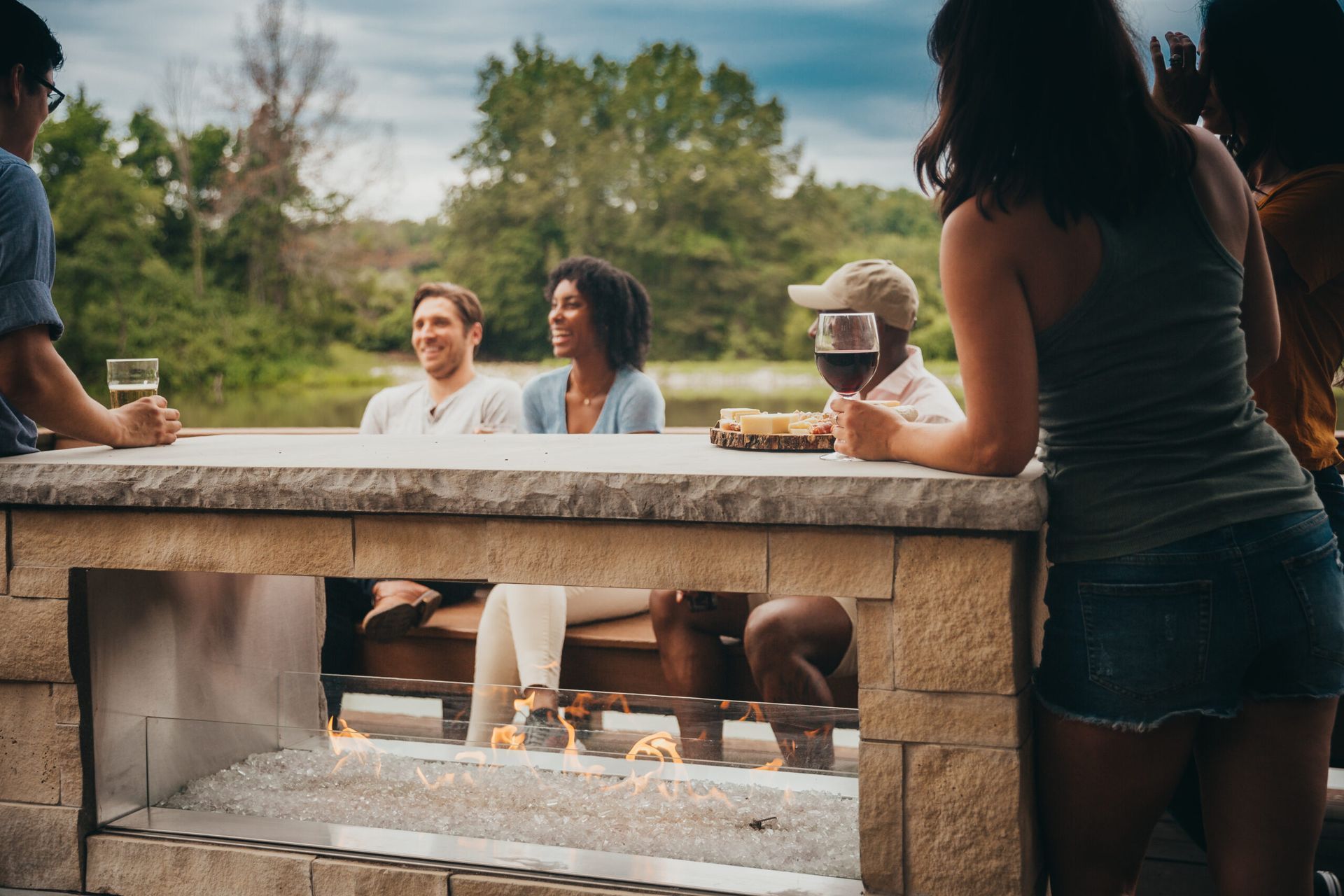 Group of people at an outdoor gathering, around a stone structure with a small fire, near water.