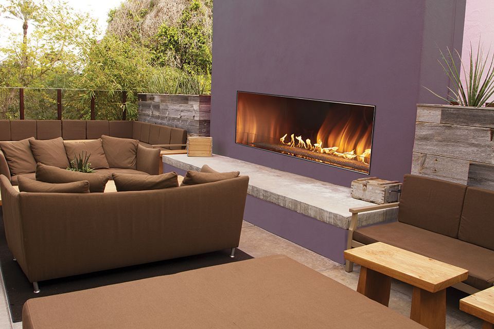 Outdoor lounge with brown furniture, fireplace with flames, purple wall, and natural setting.