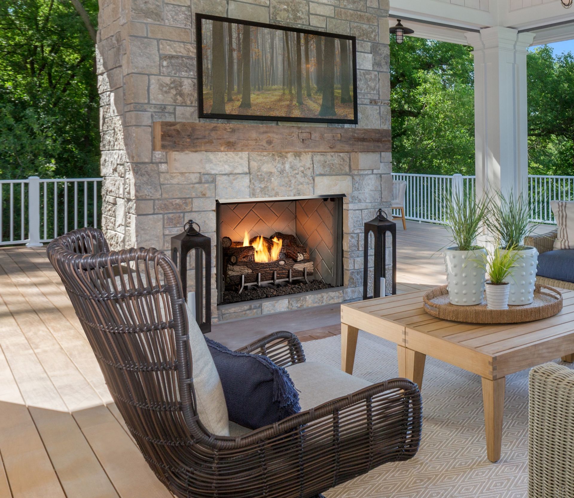 Outdoor fireplace with stone facade, TV, and wicker chairs on a deck, with green trees in the background.