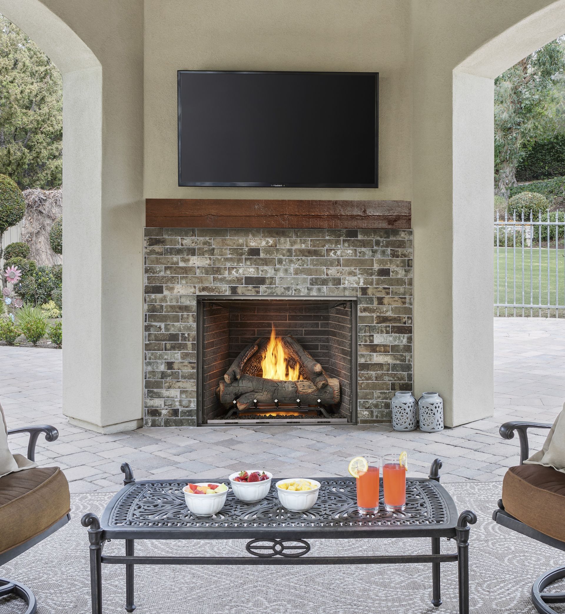 Outdoor fireplace with a mounted TV, seating area, and table with drinks and fruit.