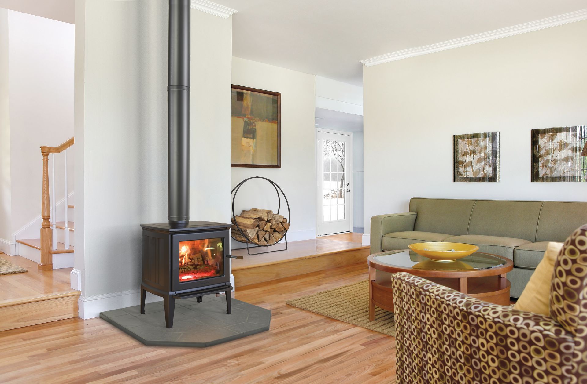 Wood-burning stove in a living room, with a lit fire, connected to a black chimney. The room features a sofa, rug, and art.