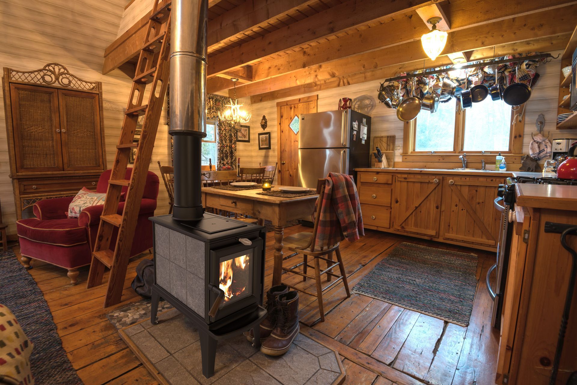 Cozy cabin interior with a wood-burning stove, kitchen, wooden beams, and a ladder to a loft area.