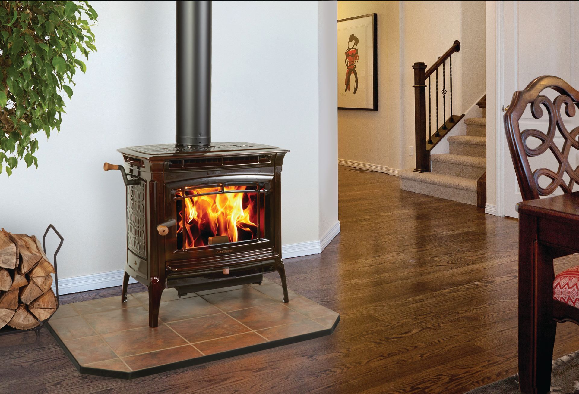 A wood-burning stove with flames in a home; logs in a basket, hardwood floor and staircase in the background.