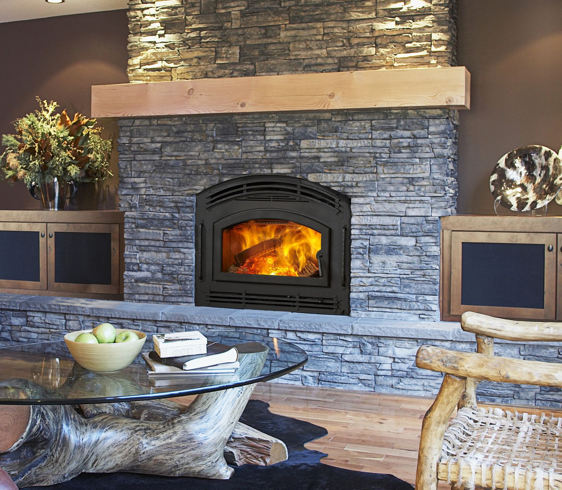 Living room with fireplace, glass table, wood floors, and large windows overlooking greenery.