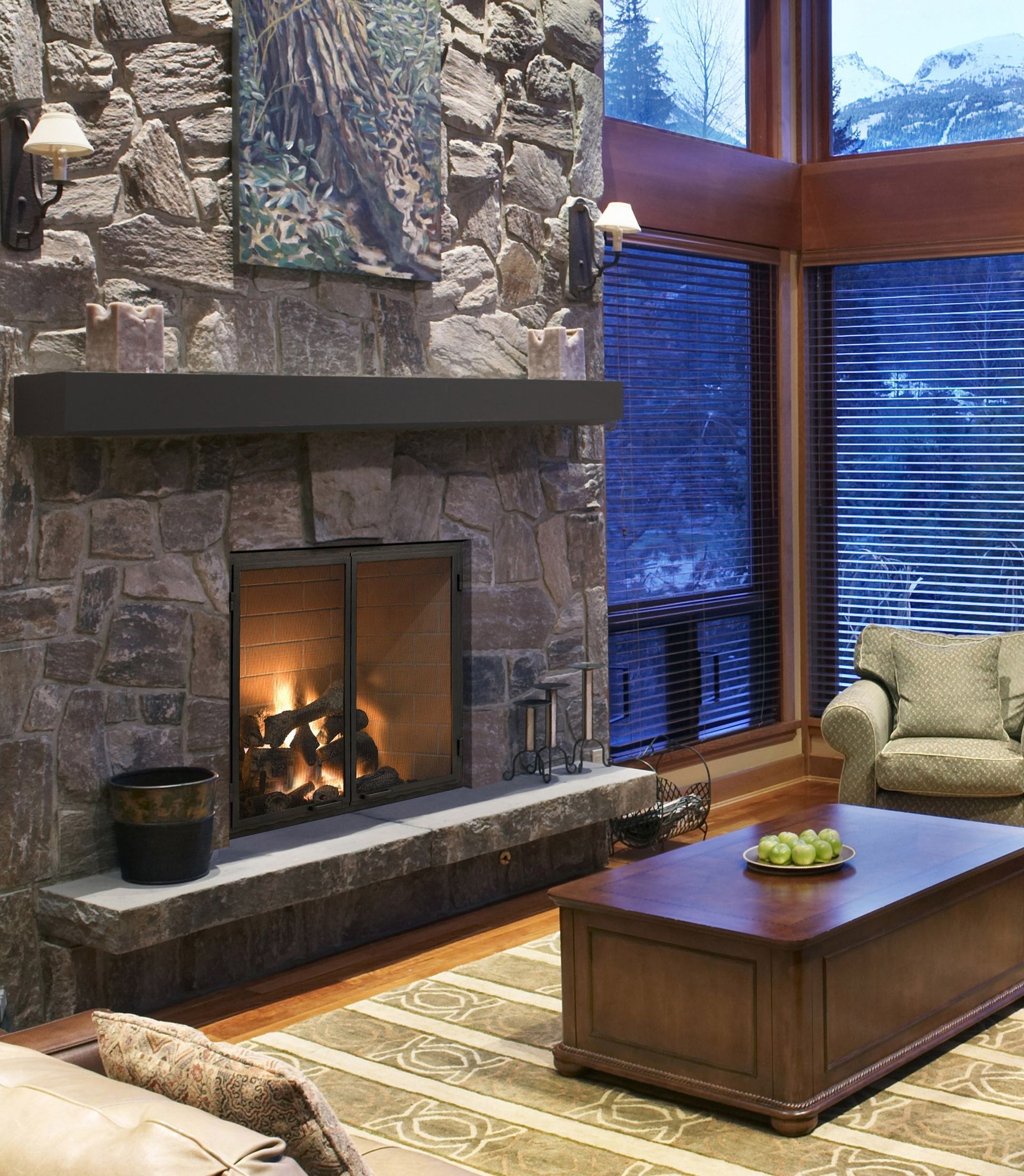 Cozy living room with stone fireplace, fire burning, dark wood mantel, large window, and brown coffee table.