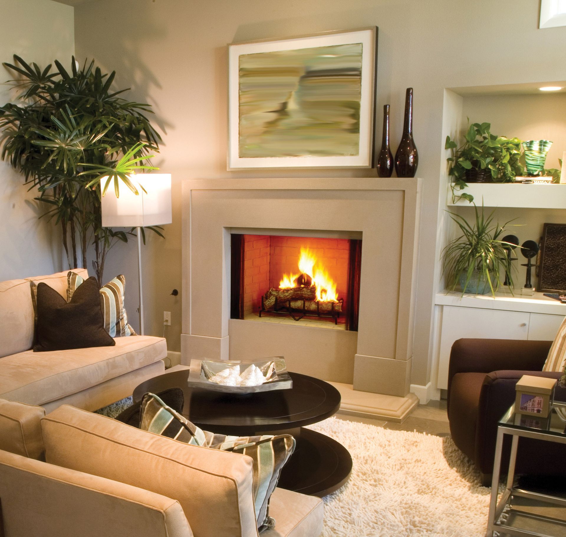 Cozy living room with fireplace, beige couch, round black coffee table, artwork, plants, and a brown armchair.