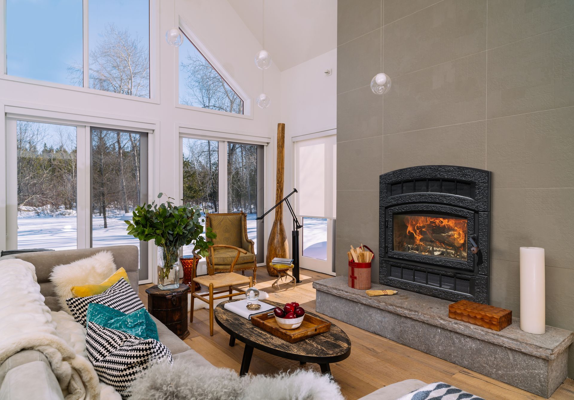 Living room with fireplace, windows overlooking snowy landscape. Gray walls, light wood floor, neutral-toned furniture.