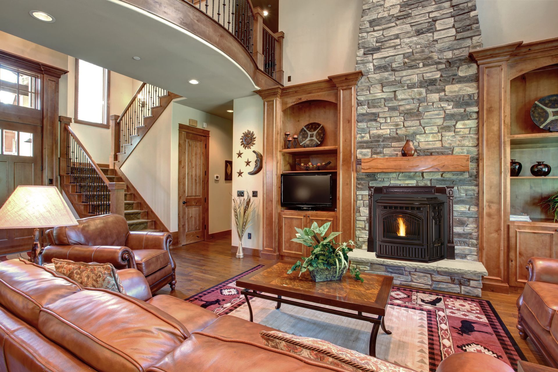 Cozy living room with fireplace, stone wall, built-in shelving, brown leather sofas, and wooden staircase.
