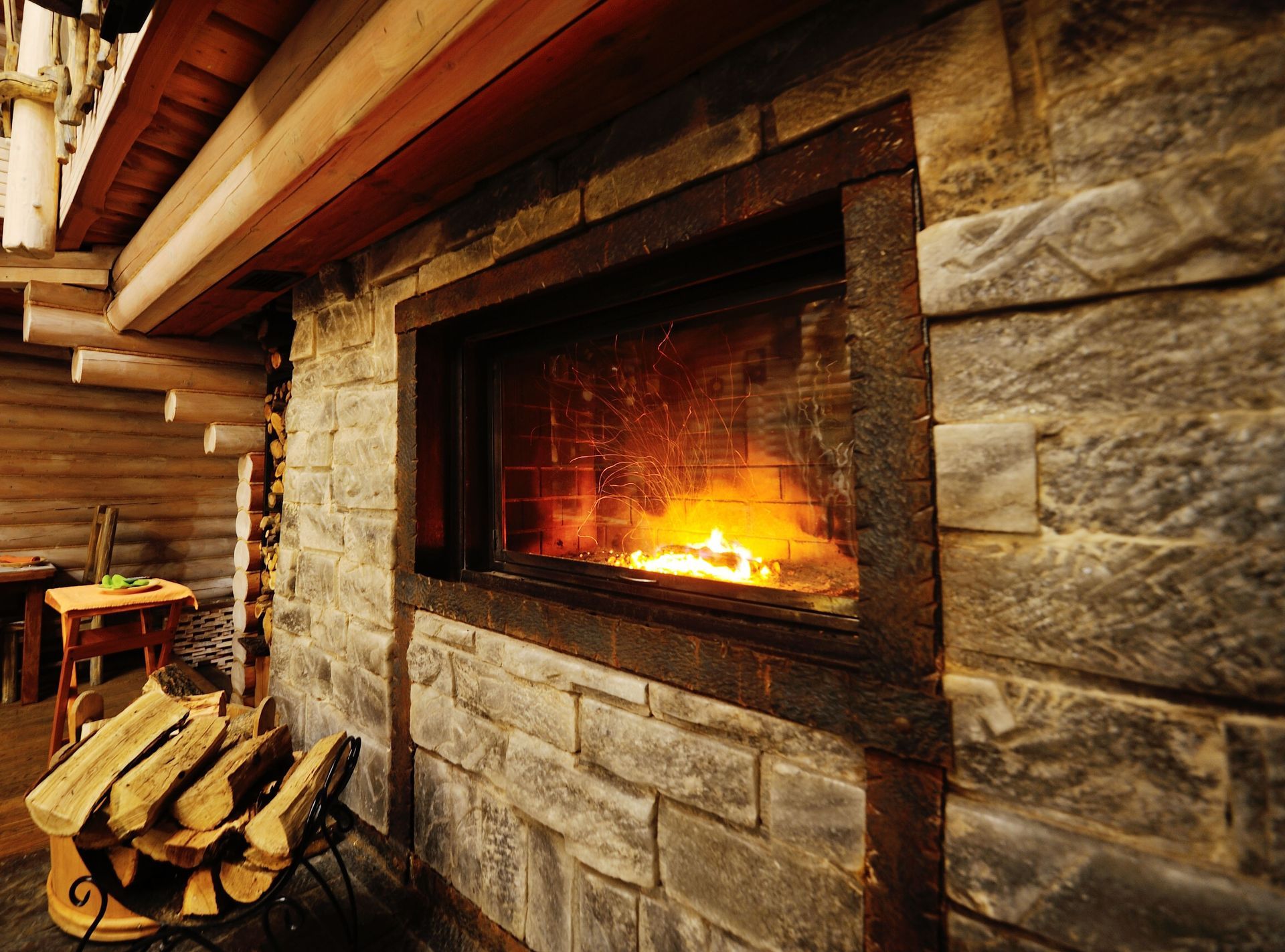Stone fireplace with a burning fire, firewood beside it, inside a log cabin.
