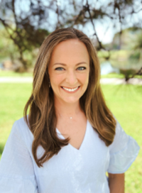 Woman with brown hair smiles outdoors, wearing a white top, trees and water blurred in the background.