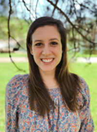 Woman with long brown hair smiles, wearing a floral print top, outdoors.