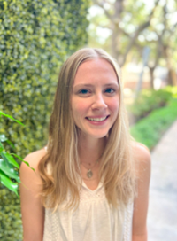 Blonde woman smiles at the camera, wearing a white top, standing in front of greenery with trees blurred in the background.