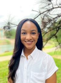 Woman with long, dark hair smiles, wearing a white shirt outdoors near greenery and a pond.