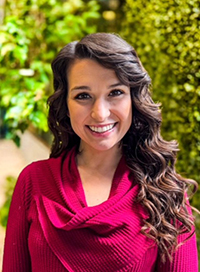 Woman with brown hair smiling in a red sweater against a green plant backdrop.