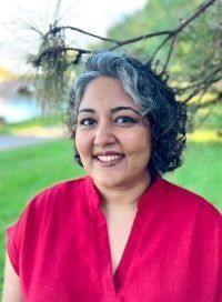 Woman with graying dark hair smiles outdoors, wearing red shirt, set in a park.
