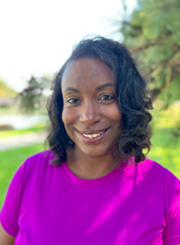 Woman with dark hair smiling, wearing a purple shirt, outdoors with trees and grass.