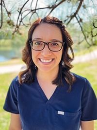 Woman in blue scrubs smiling, wearing glasses, outdoors.