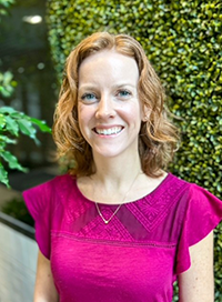 Woman with red hair smiles, wearing a magenta top, in front of a green plant wall.