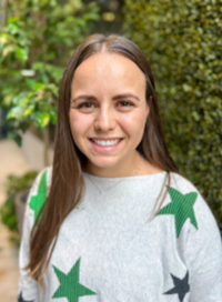 Woman smiling, wearing gray sweater with green and black star designs, outdoors with green foliage.