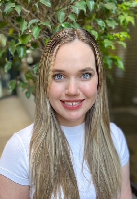 Woman with blonde hair smiling in front of leafy green background. Wearing a white shirt.
