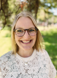 Woman with glasses, smiling, in front of green trees. She is wearing a white lacy sweater.
