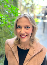 Blonde woman smiling, wearing a beige vest and black shirt outdoors, in front of greenery.
