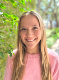Blonde woman with a big smile, wearing a pink sweater, stands outside near green foliage.