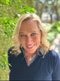Blonde woman smiling, wearing a navy top, outside with foliage in background.