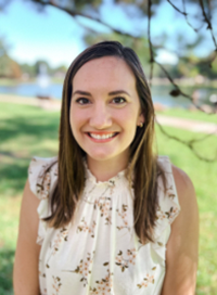 Woman with brown hair smiles outdoors, wearing a floral top. A park with a fountain is in the background.