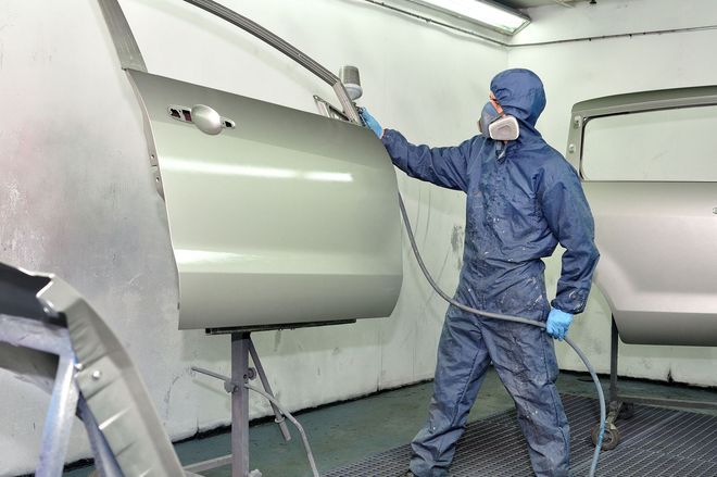 Worker in blue protective suit spray-painting a car part inside a paint booth
