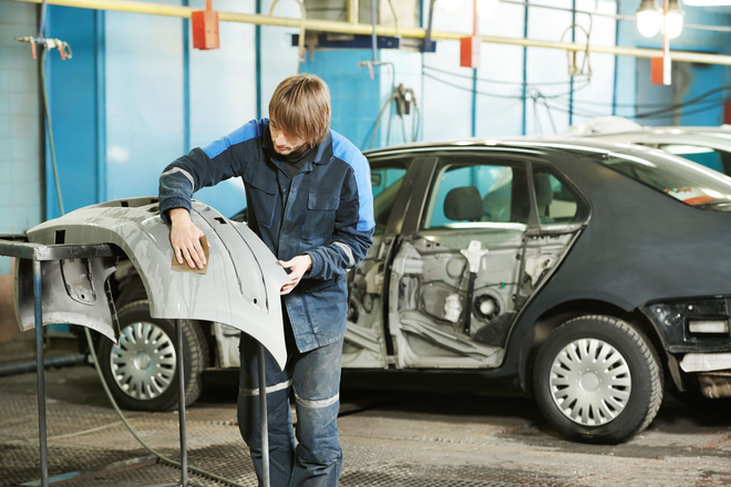 Mechanic examining a detached car door beside a damaged black sedan in a repair shop