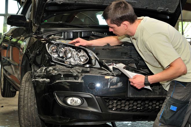 Mechanic inspecting damaged black car front with hood open in a repair garage