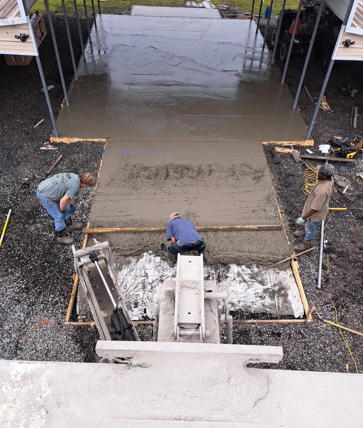 Construction workers pouring and leveling wet concrete for a driveway under a carport.