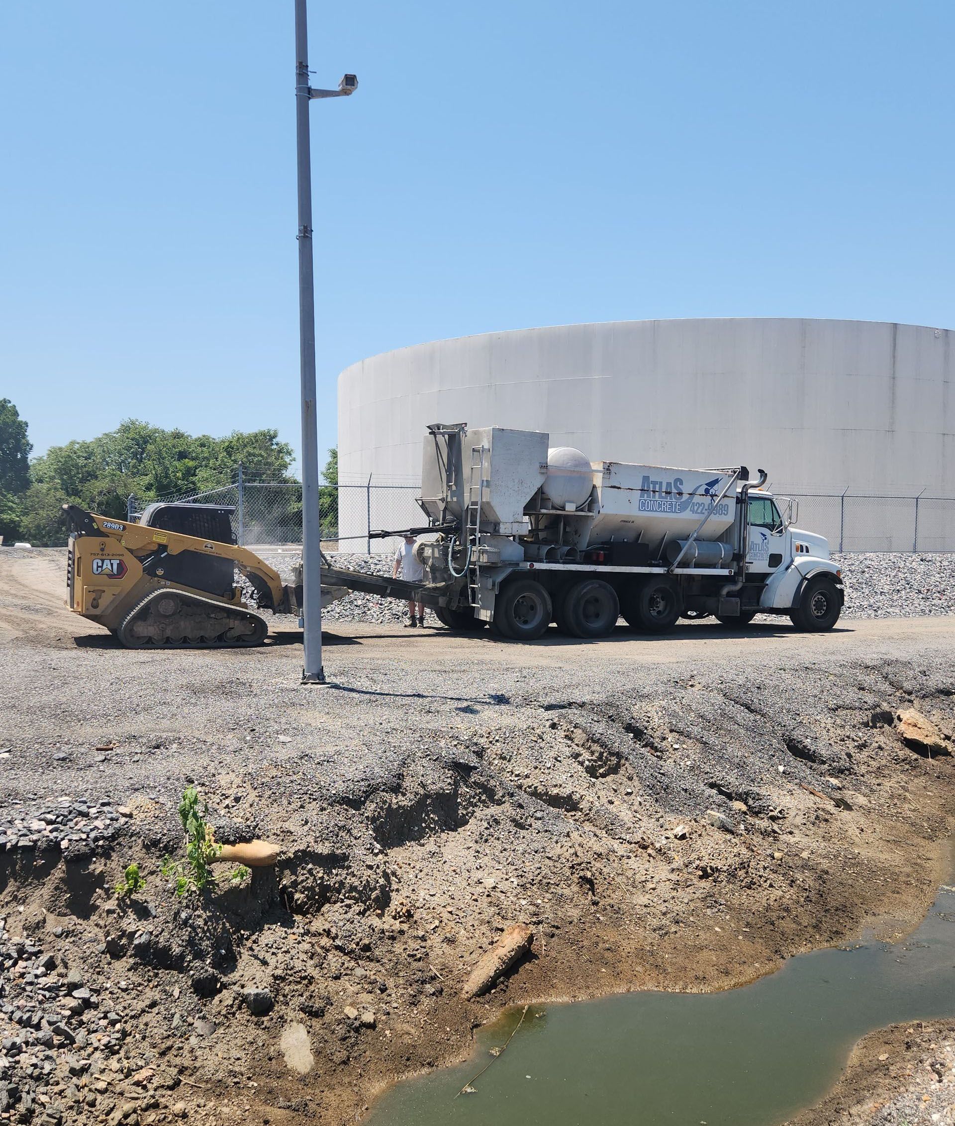A large concrete truck with a milling machine attachment is parked on a gravel lot near a white industrial tank.