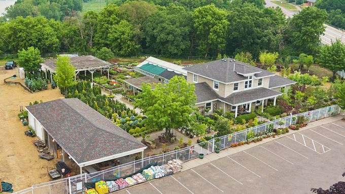 Aerial view of a nursery and garden center featuring a house, greenhouses, outdoor plant displays, and a parking lot.
