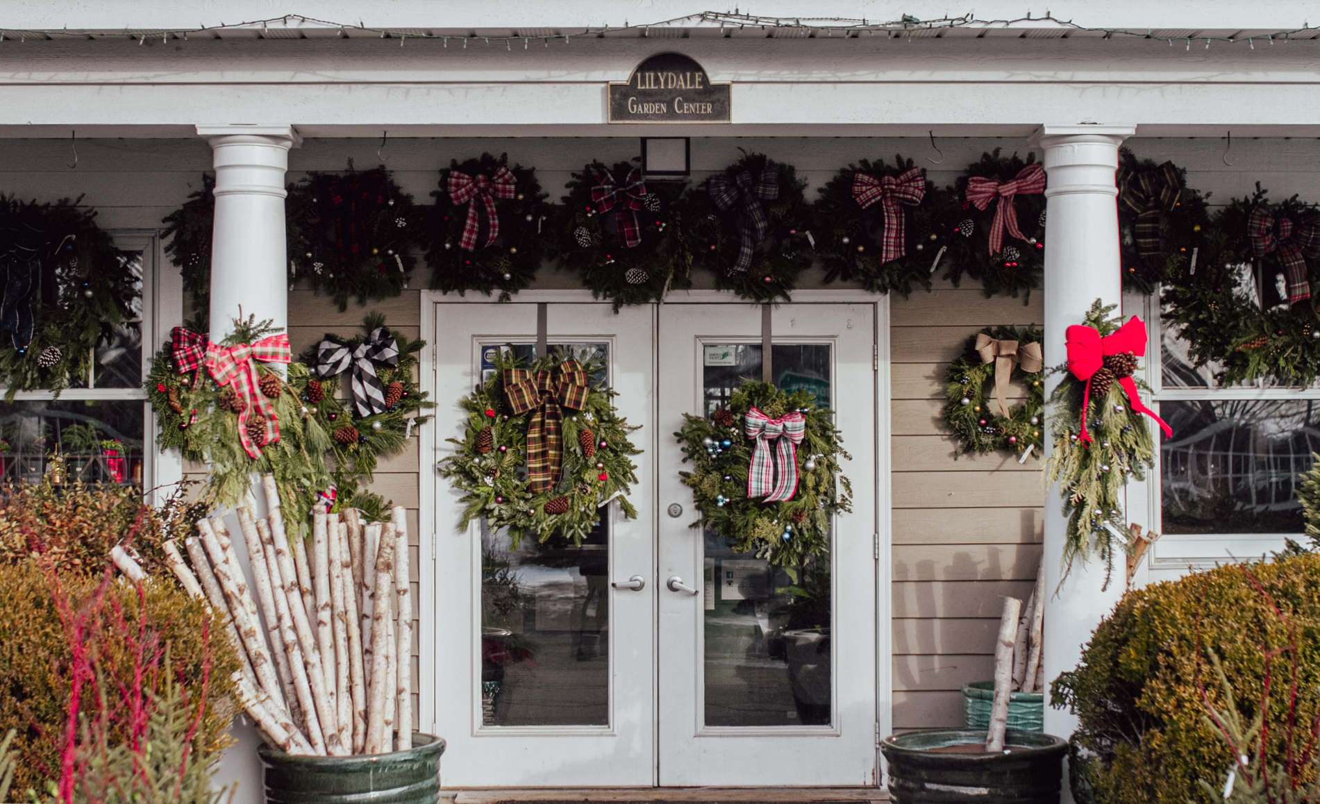 A building entrance decorated with festive Christmas wreaths and bows, featuring white pillars and stacks of birch logs.