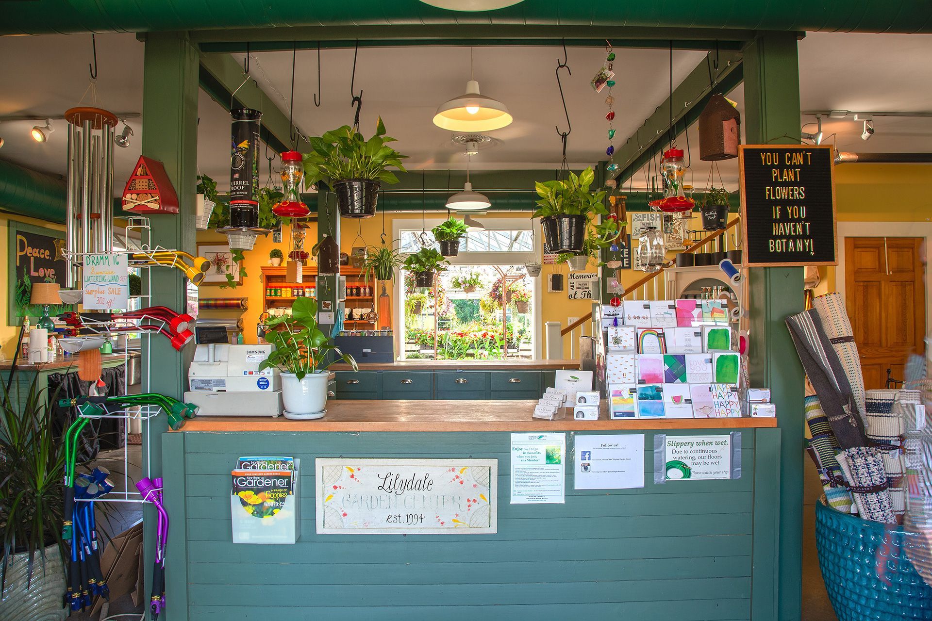 A view of a green service counter in a garden shop, with hanging plants, wind chimes, greeting cards, and a chalkboard sign.