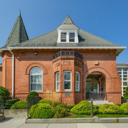 Red brick Victorian-style house with a turret, arched windows, and trimmed hedges under a blue sky