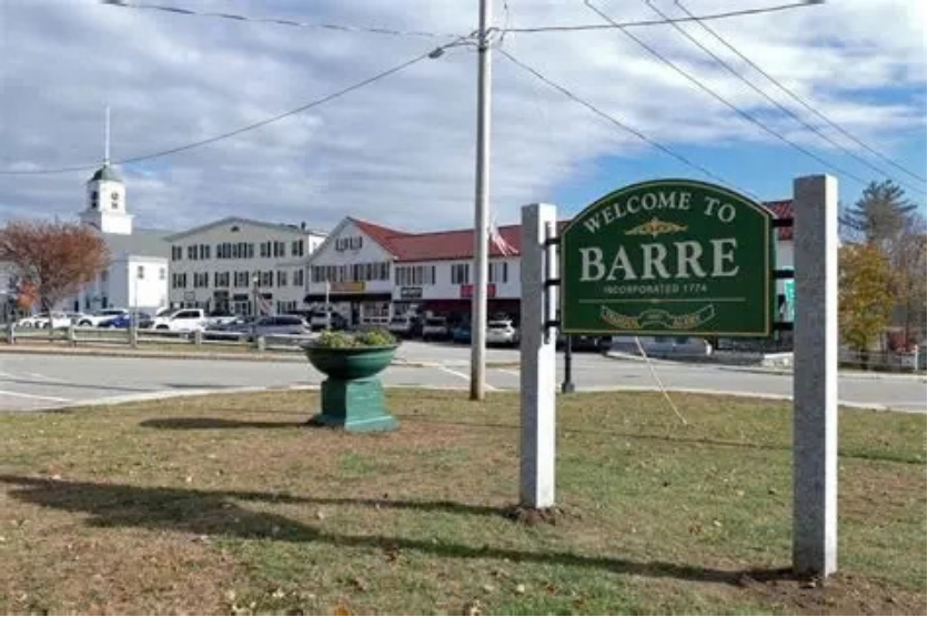 Welcome to Barre roadside sign beside town buildings and a green planter under cloudy skies