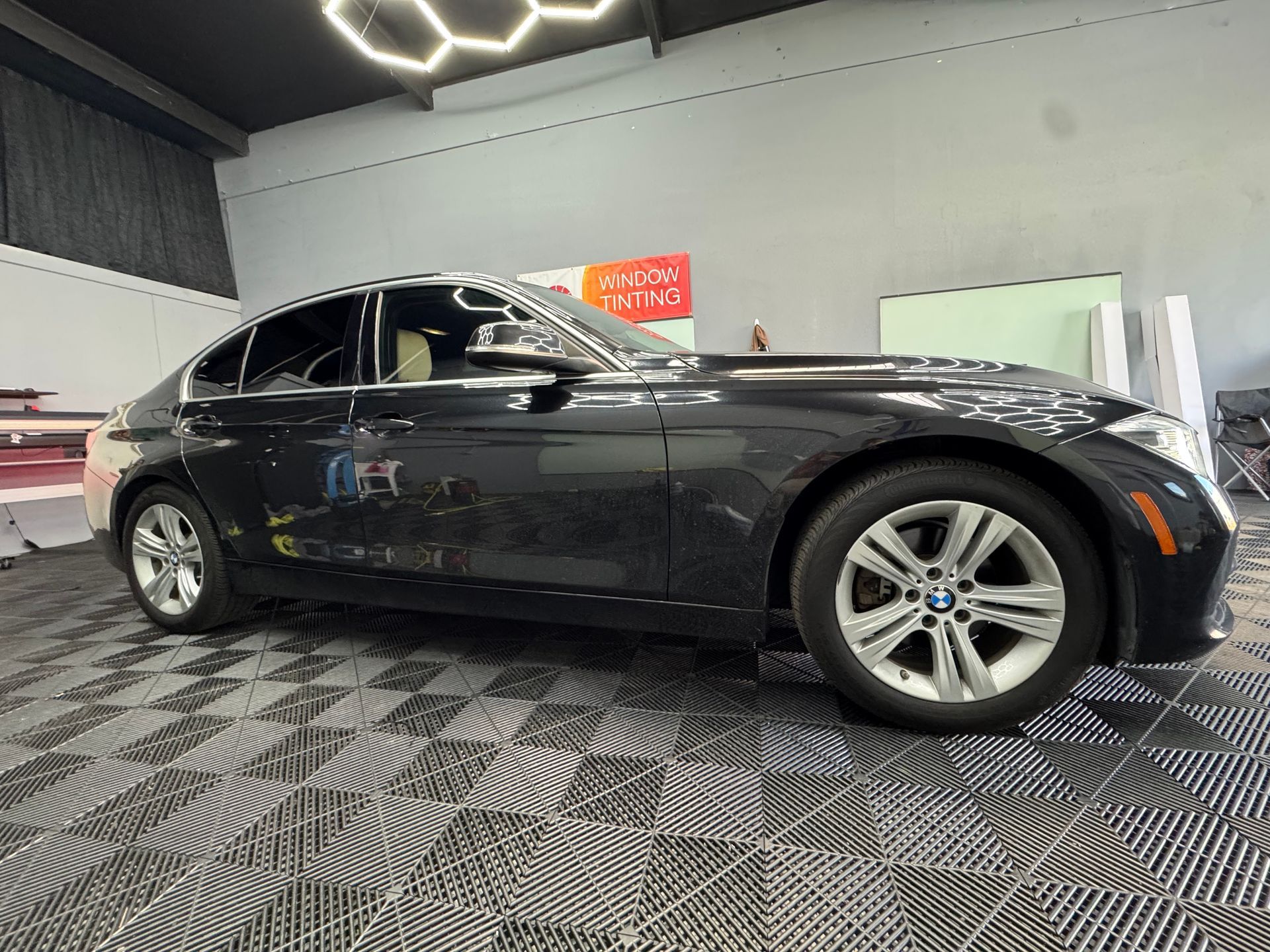 Black BMW sedan in a garage, tinted windows, silver rims, on a patterned floor.
