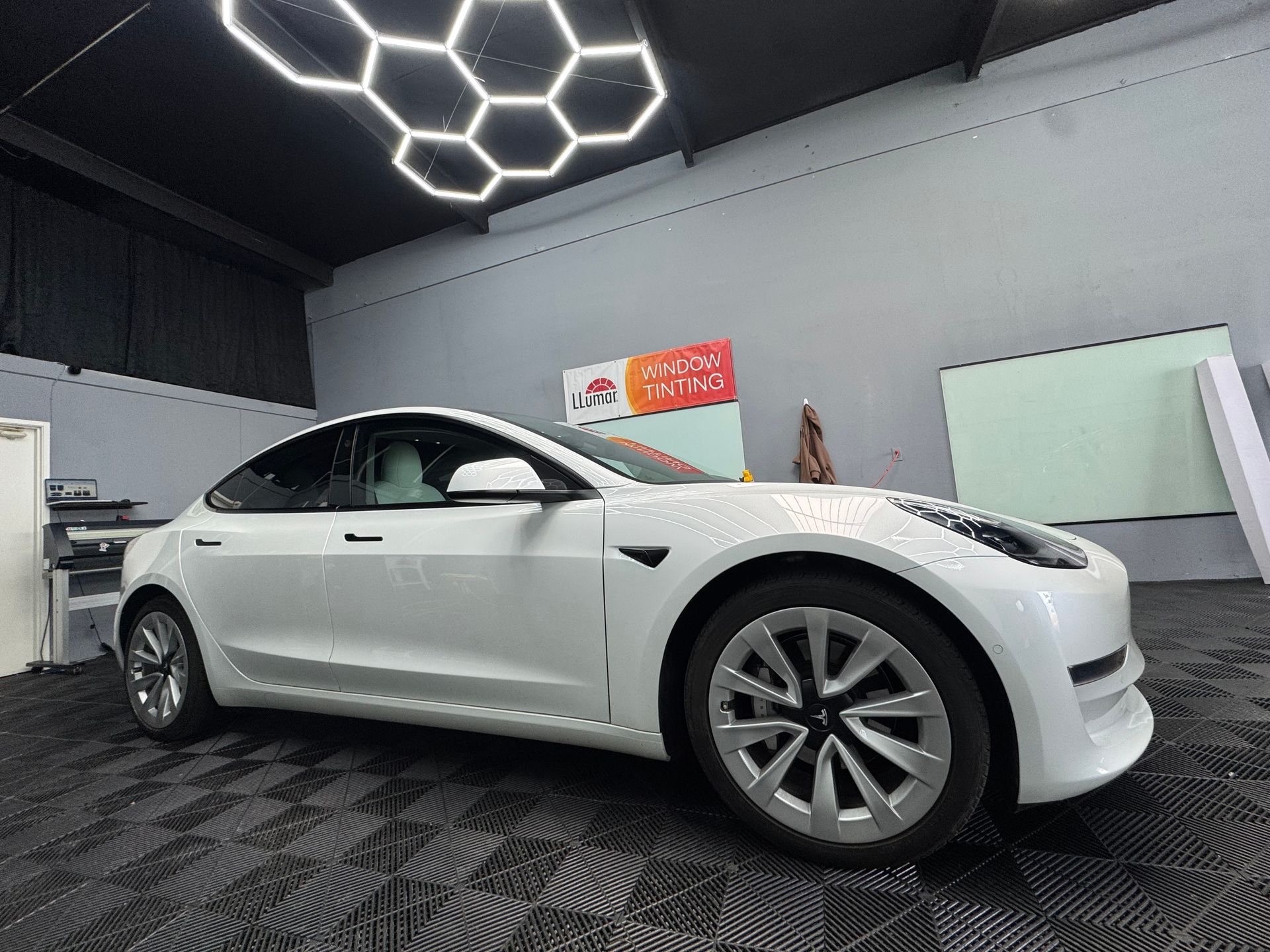 White Tesla Model 3 in a well-lit garage, with geometric ceiling lights and black tiled floor.