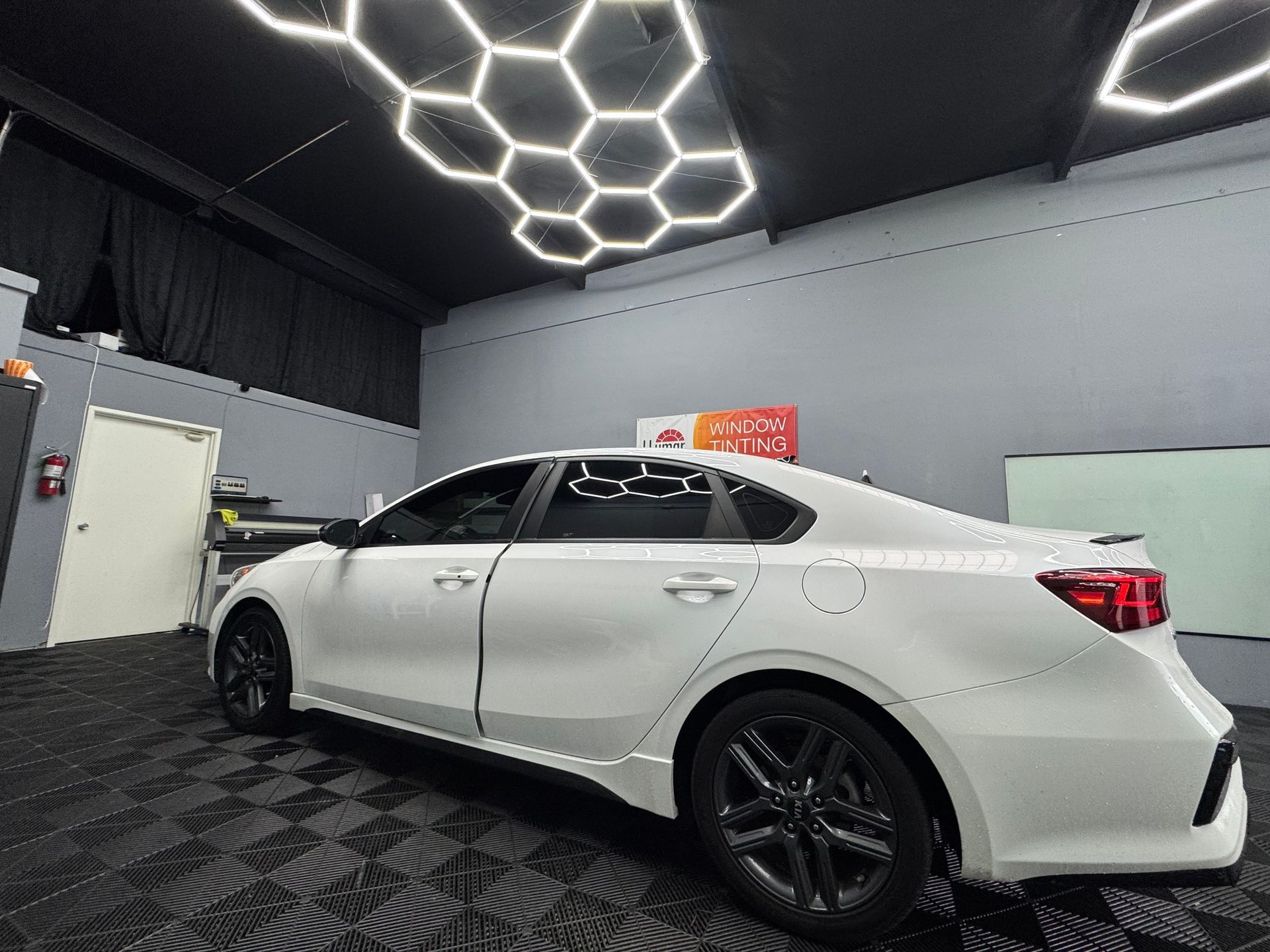 White sedan parked in a garage with black trim and wheels. Hexagon lights on the ceiling.