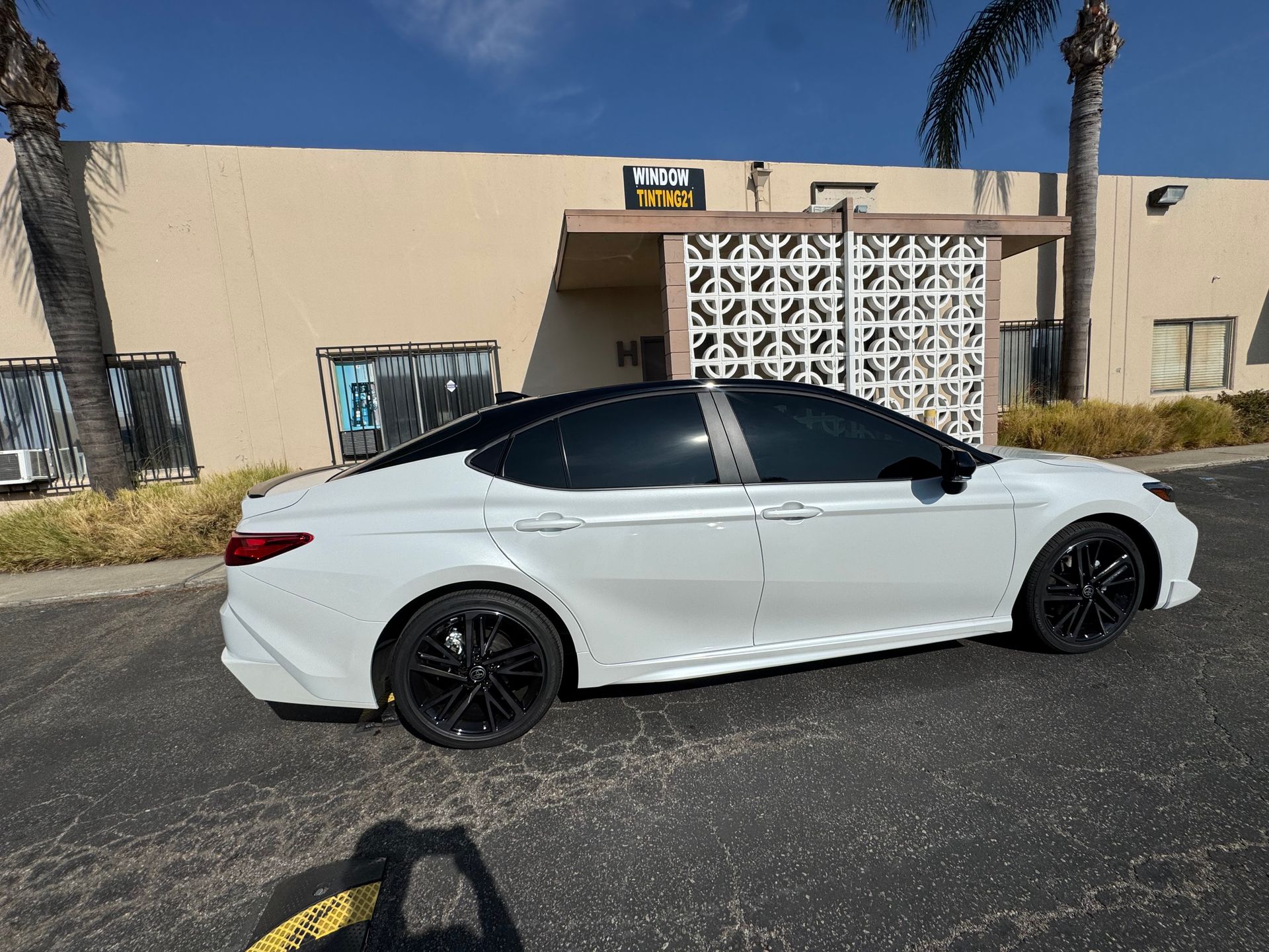 White Toyota Camry with tinted windows and black rims parked in front of a building on a sunny day.