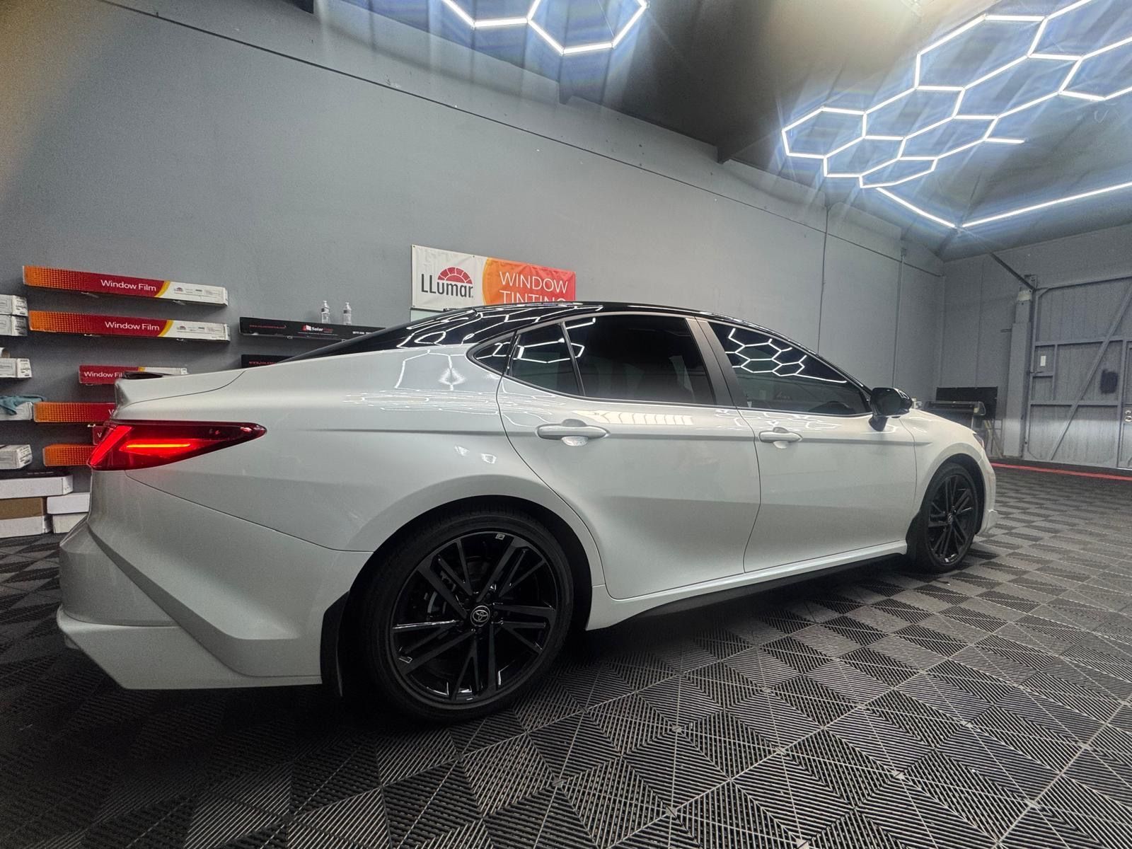 White Toyota Camry with black wheels inside a well-lit garage, with decorative hexagonal lights.