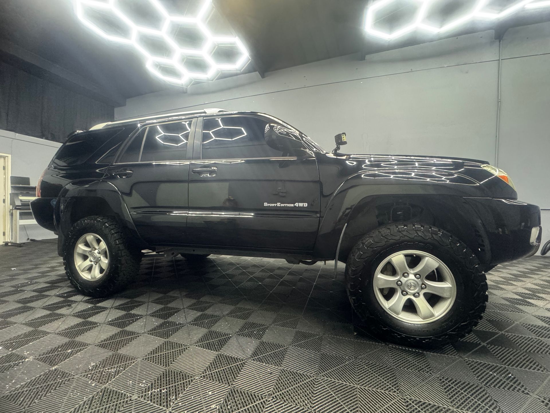 Black Toyota 4Runner SUV, lifted with off-road tires, parked inside a garage with hexagon lights and checkered floor.