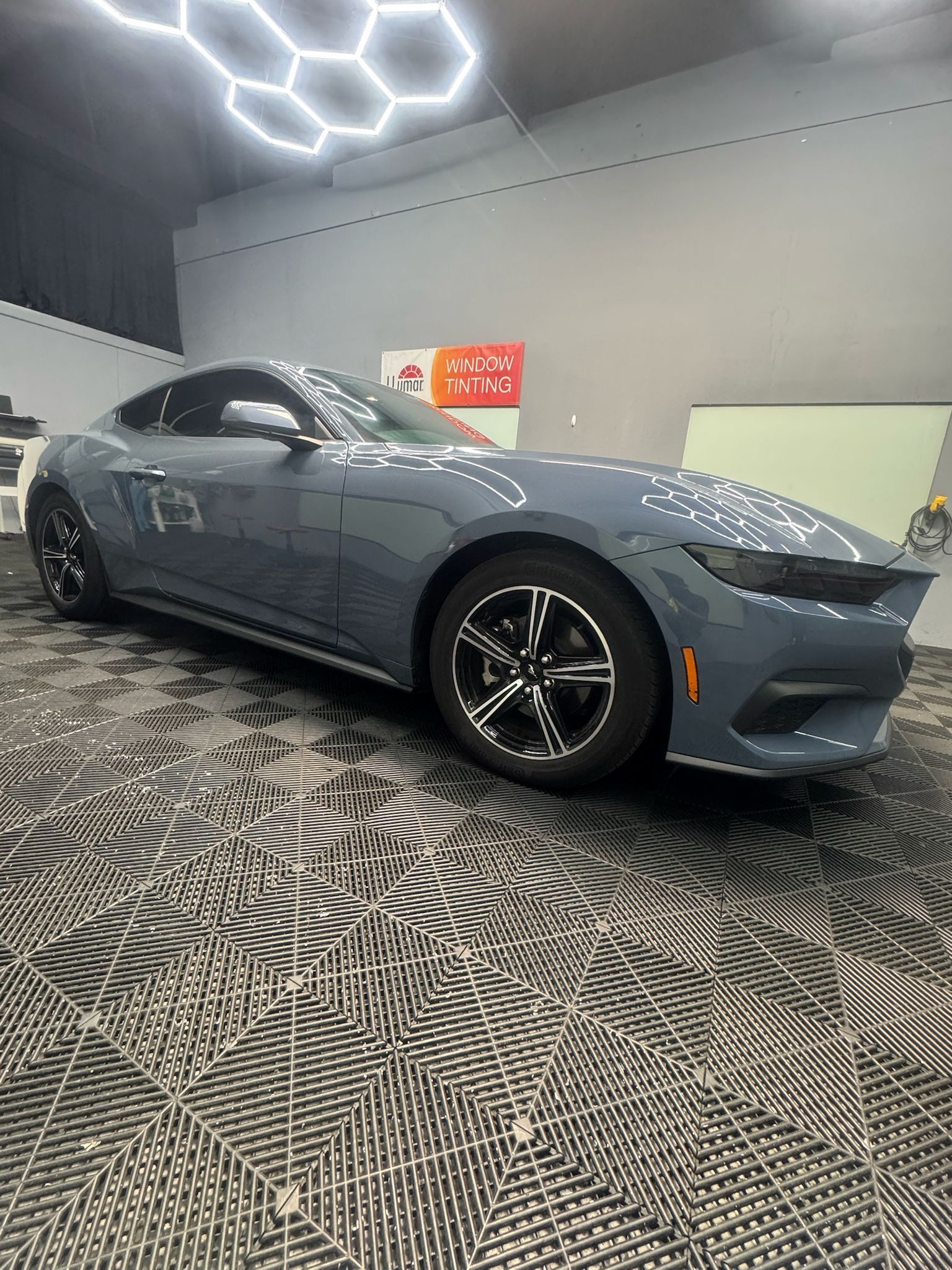 Gray sports car on patterned floor, under hexagonal lights.