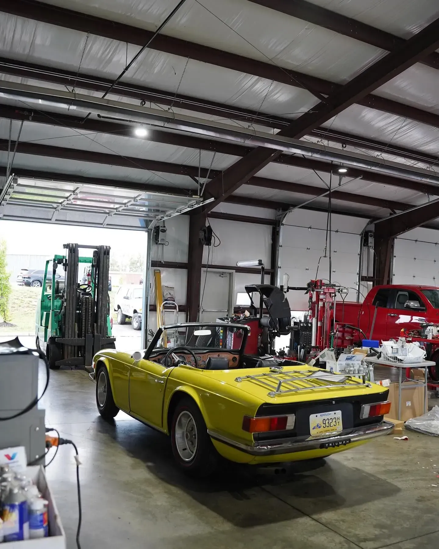 Yellow classic car in a garage with a forklift, red truck, and open door.