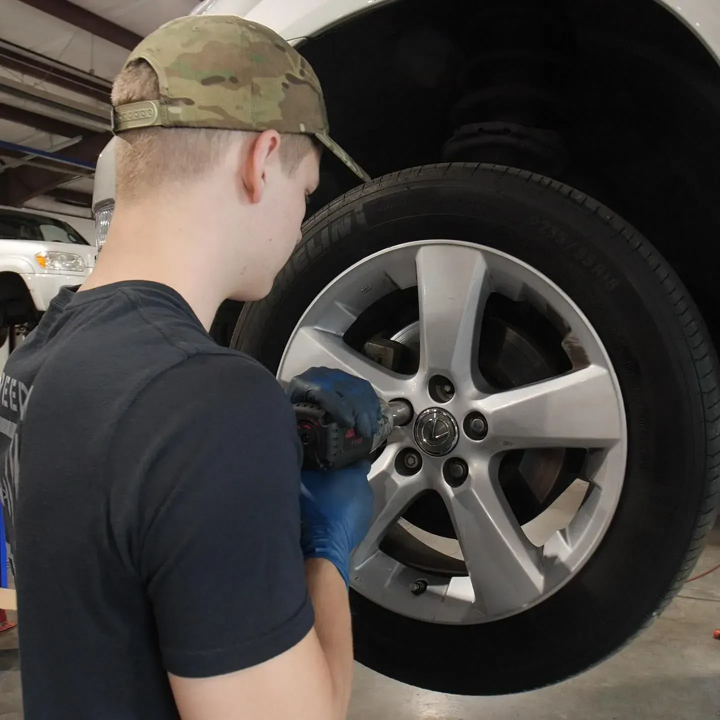A person in a camouflage cap and blue gloves removing a car wheel in a garage.