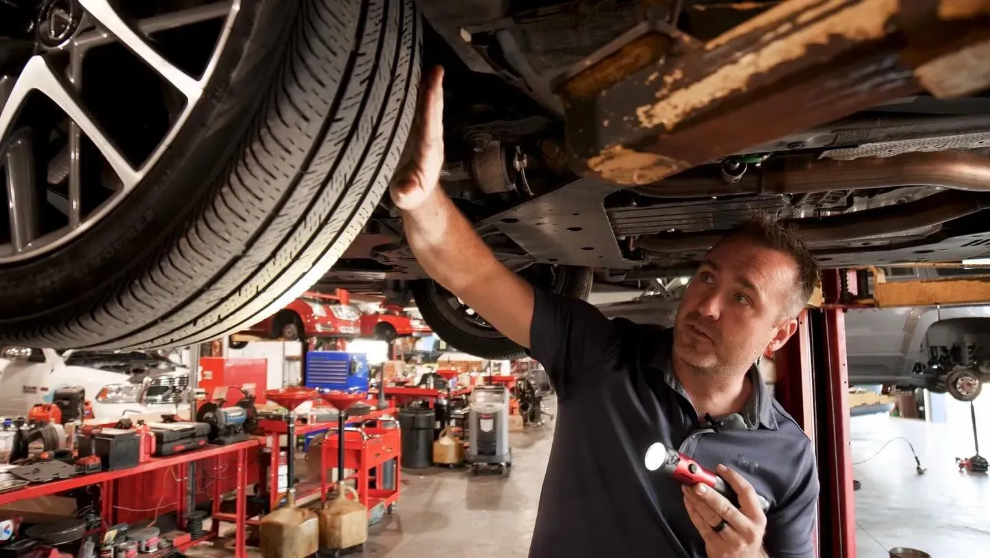 Mechanic inspecting undercarriage of a car in a shop. He uses a flashlight, pointing and explaining.